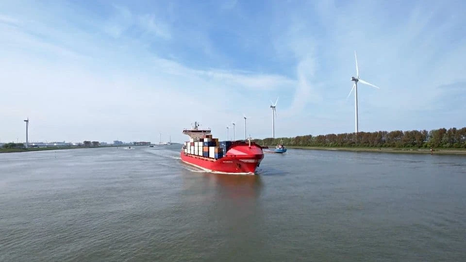 Container vessel sailing on a river with wind turbines along the shore.