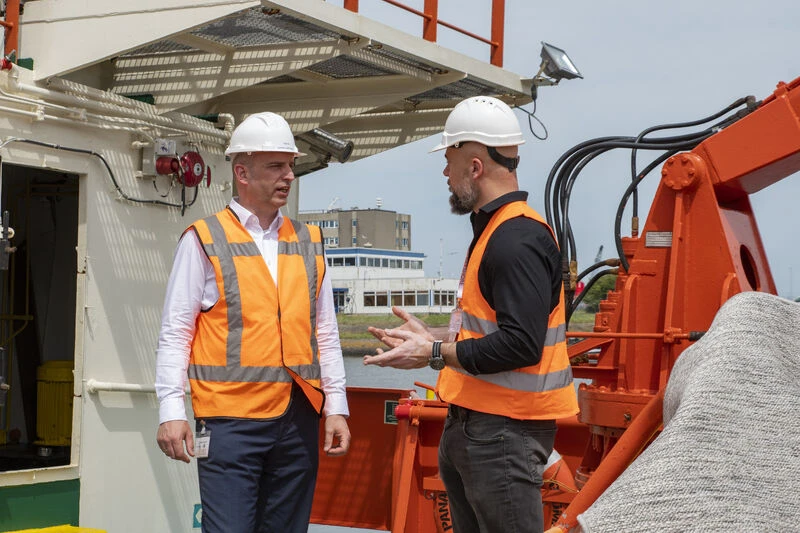 1780561271439 Two workers wearing white hard hats and high visibility orange safety vests stand on the deck of an industrial vessel, engaged in conversation beside large orange maritime equipment. Buildings and a waterfront can be seen in the background.
